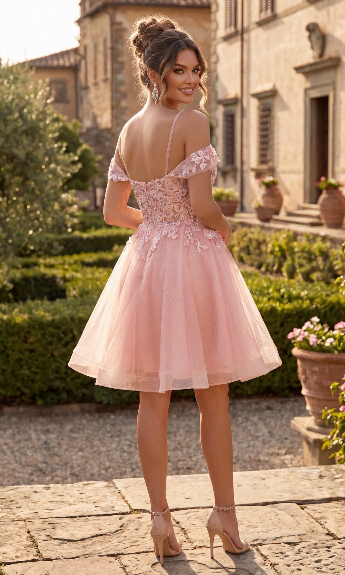 Wearing the Chicas C359 short homecoming dress, a woman stands outdoors on a stone path, smiling as she looks back over her shoulder. The off-the-shoulder pink dress features floral lace appliques and a delicate corset bodice.